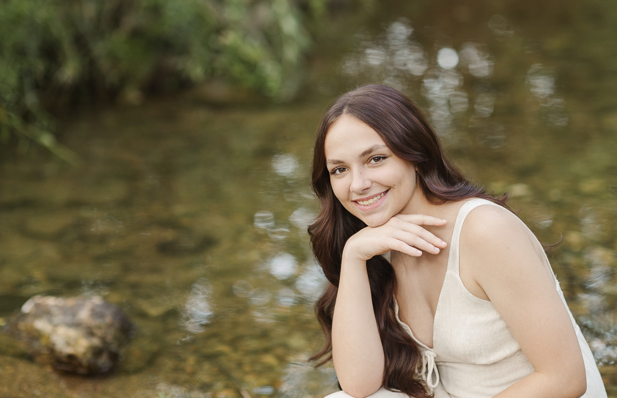 Senior girl smiling while sitting by a creek in a white dress during her portraits in York, PA.