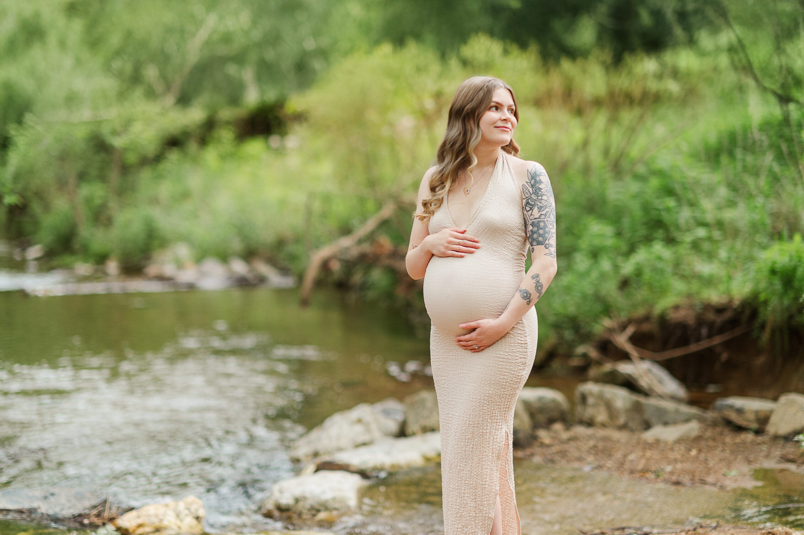 Pregnant woman in fitted tan dress holding belly near creek at golden hour, by York PA maternity photographer