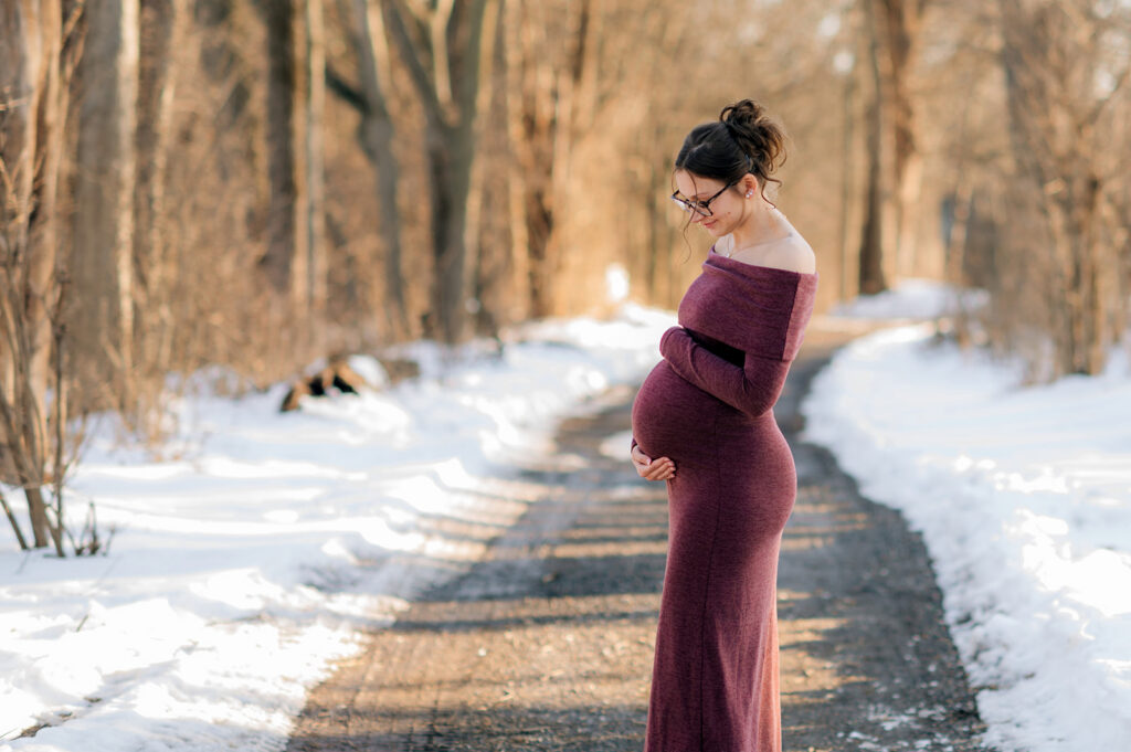 Pregnant woman in burgundy dress standing on a snow-lined trail at John Rudy Park in York, Pennsylvania.