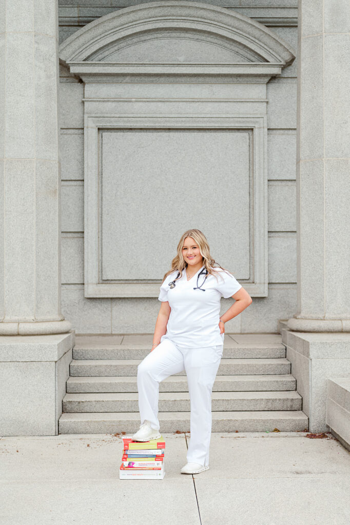 high school senior in nursing scrubs standing on books during senior photo session at Pennsylvania State Capitol