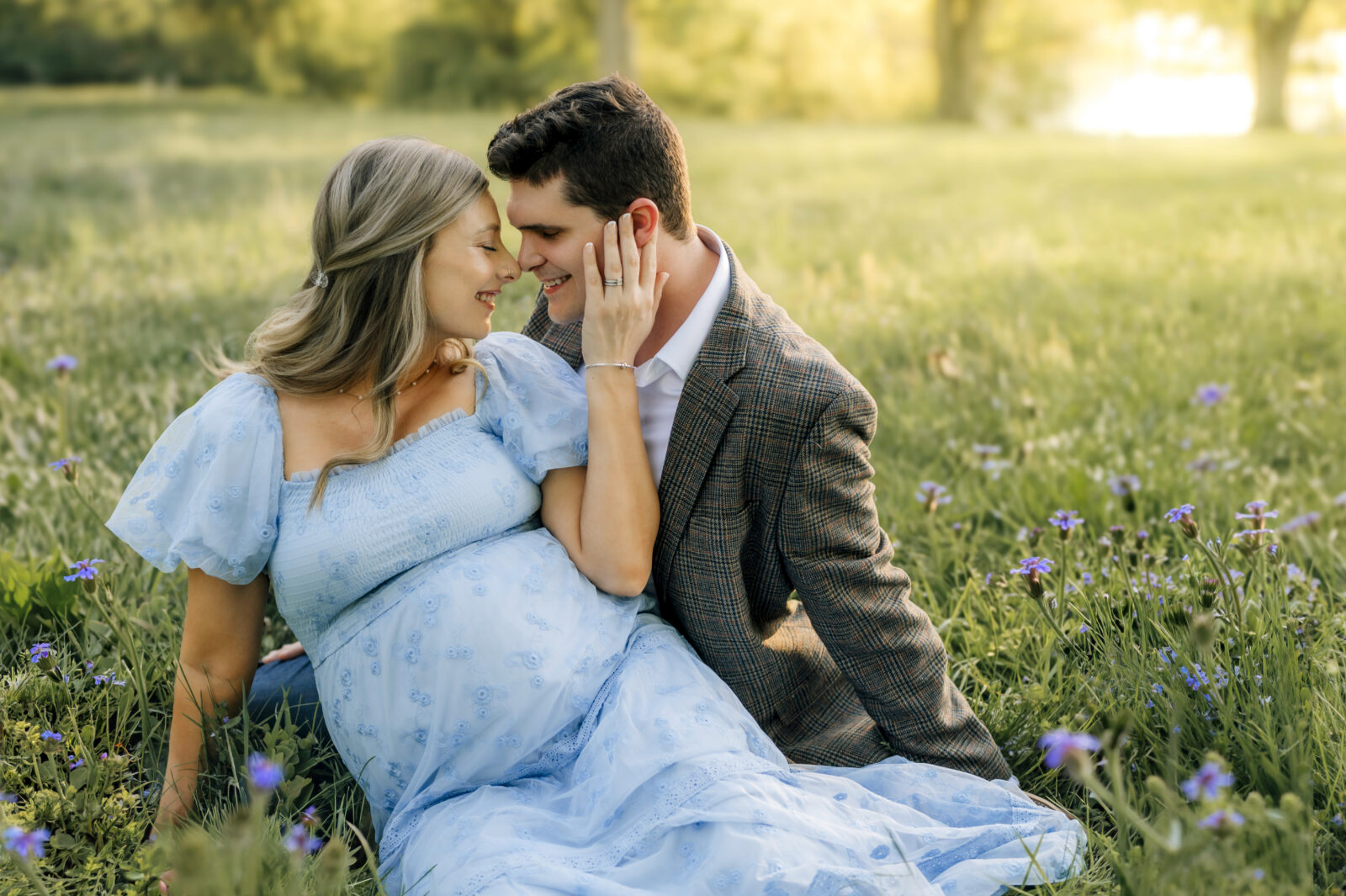 Maternity photos in York PA of a couple sitting in a field at sunset with mom in blue dress and partner leaning in close
