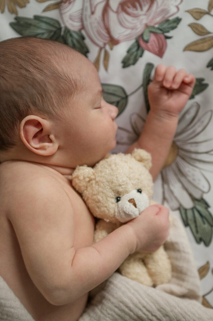 Newborn baby lying on a bed holding a small teddy bear during an in-home photography session in York, PA