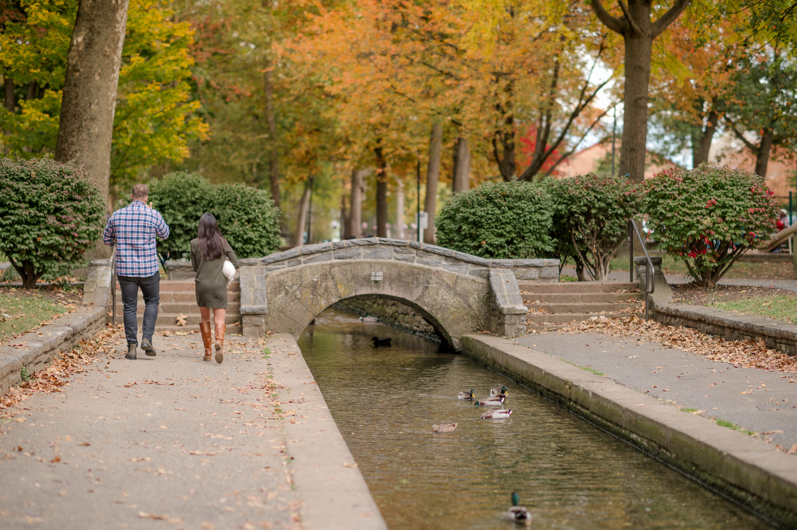 Couple walking beside the stone bridge and stream at Lititz Springs Park during a fall photography session in Lititz, PA