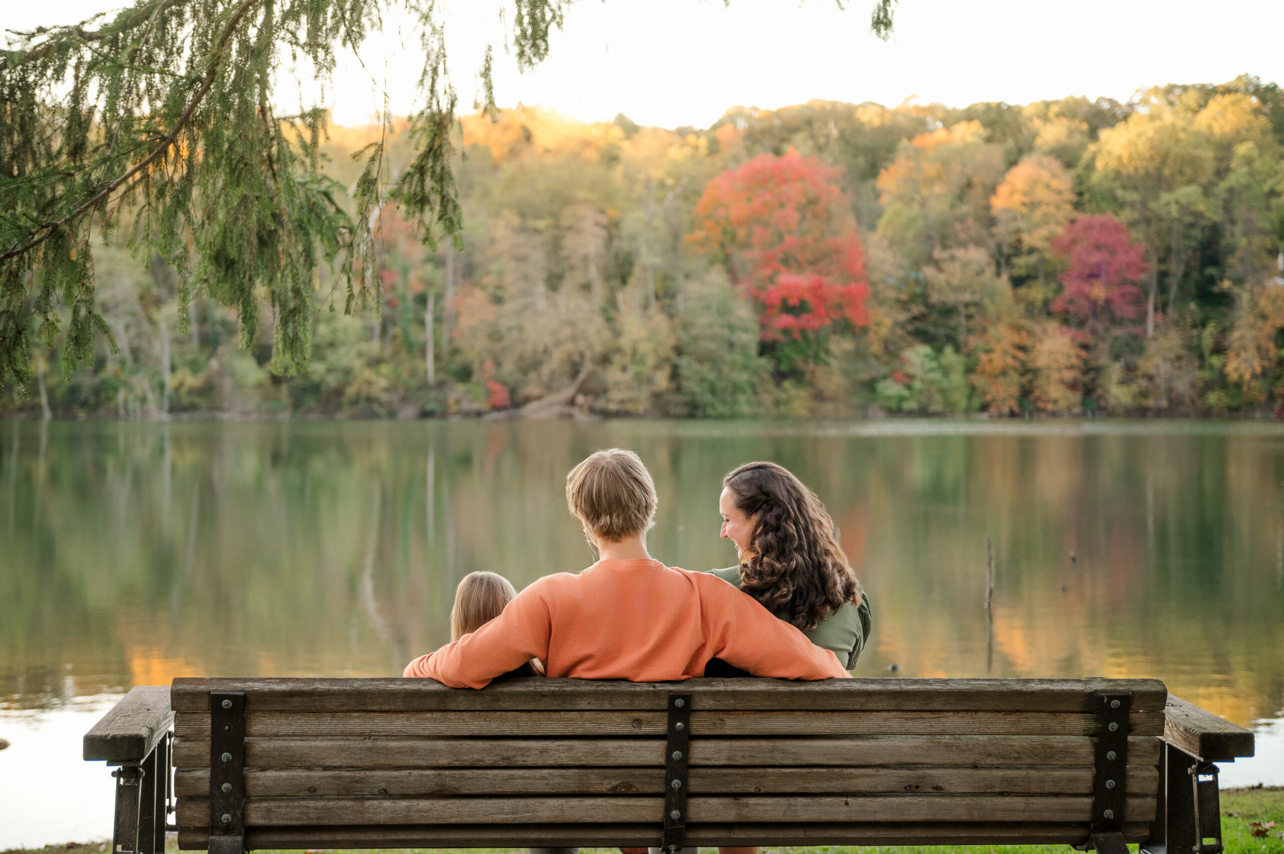 Family of three sitting together on a wooden bench overlooking a lake and colorful fall trees in Lancaster, PA.