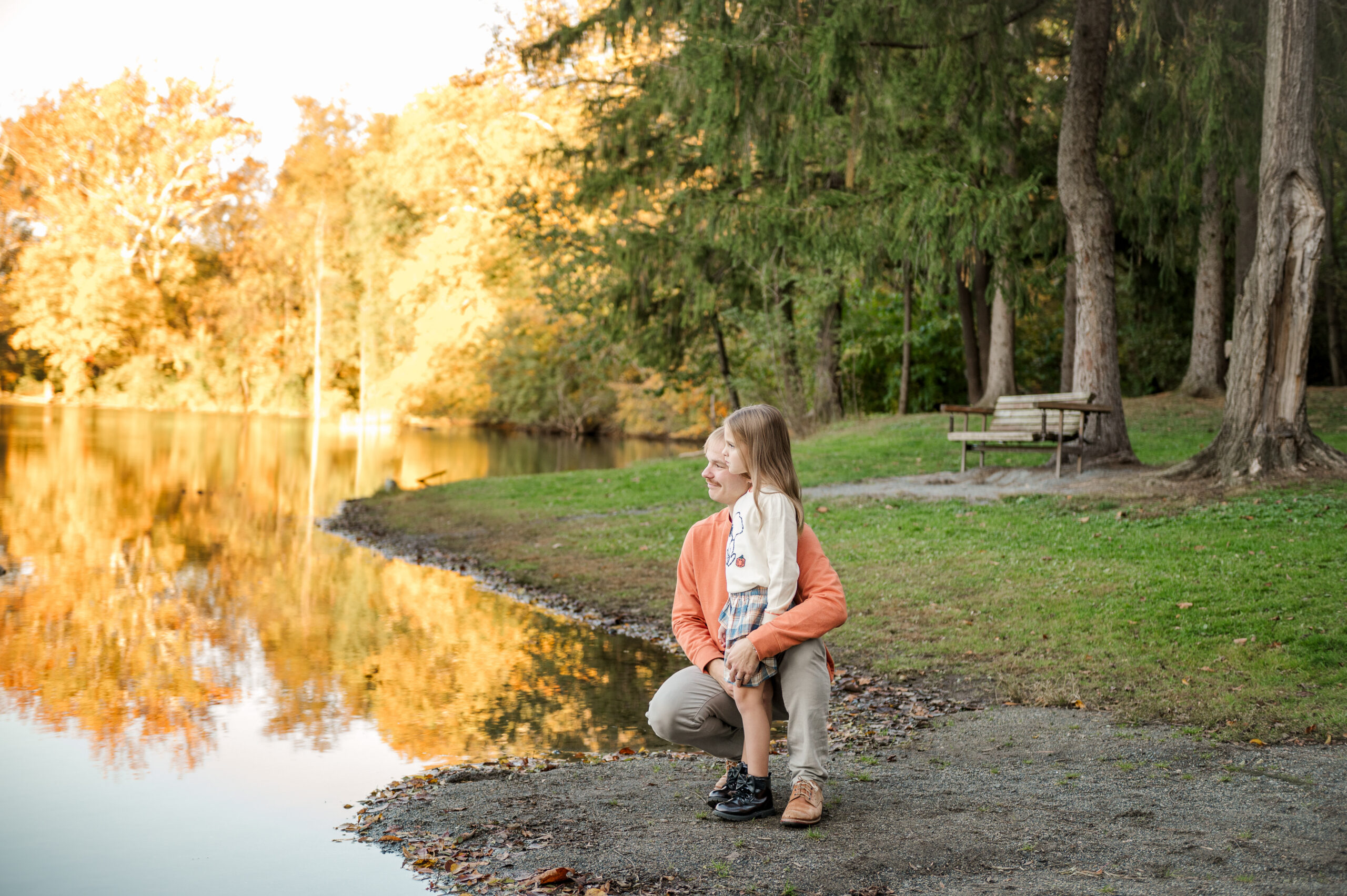 Dad holding his young daughter beside a lake surrounded by autumn trees during a fall family photo session in Lancaster, PA