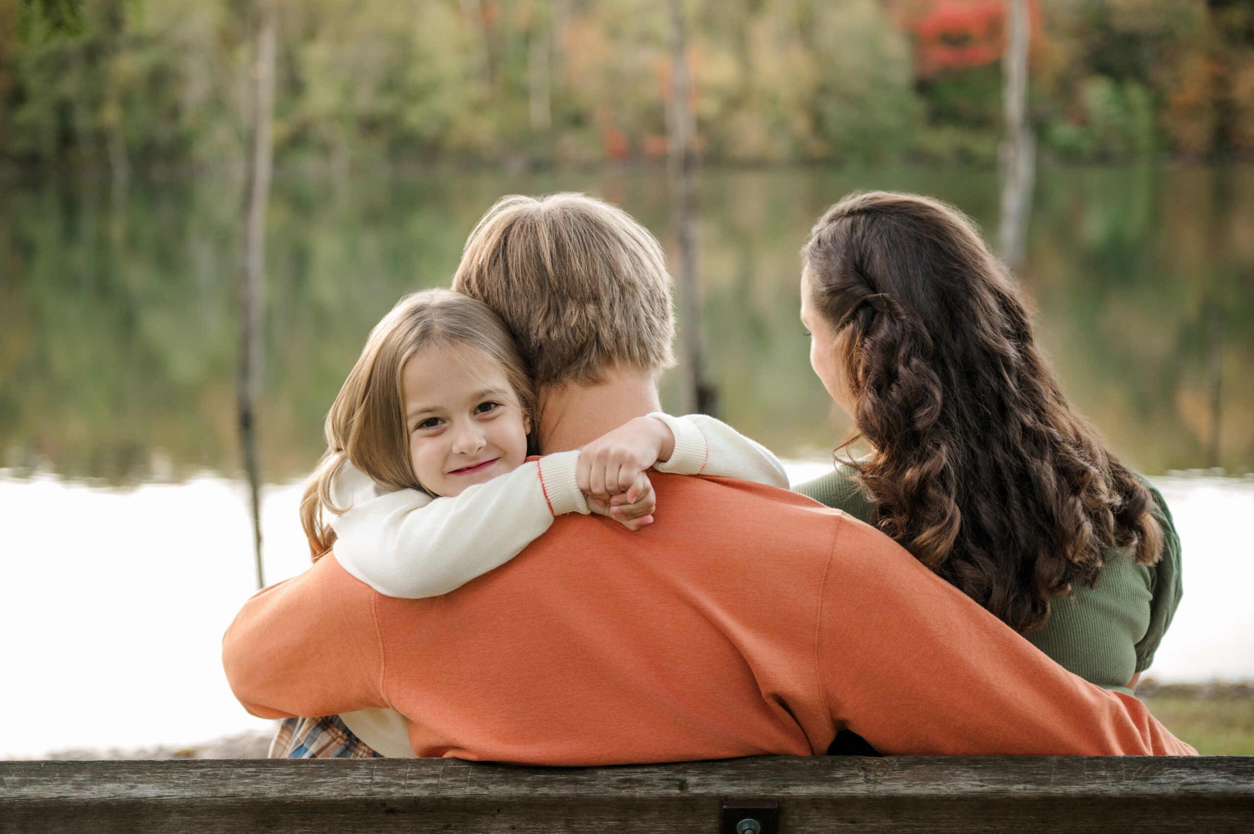 Young daughter hugging her dad while sitting with her parents beside a lake during a fall family session in Lancaster, PA