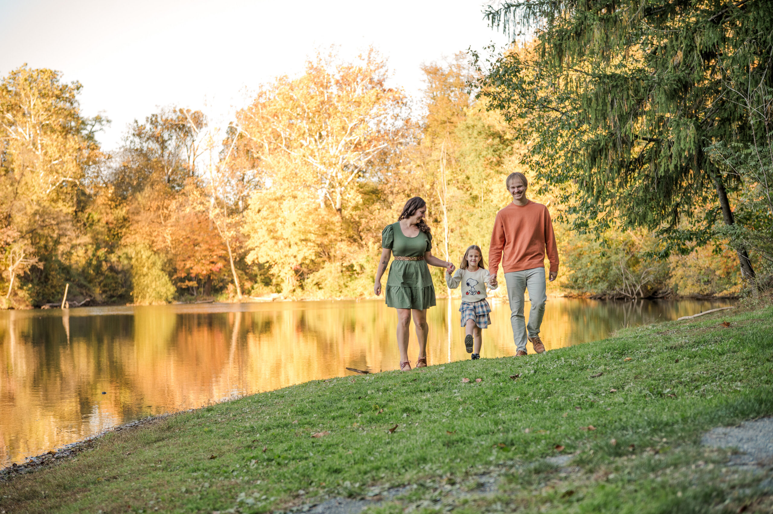 Family of three walking together beside a lake surrounded by colorful fall foliage during a family photography session in Lancaster, PA