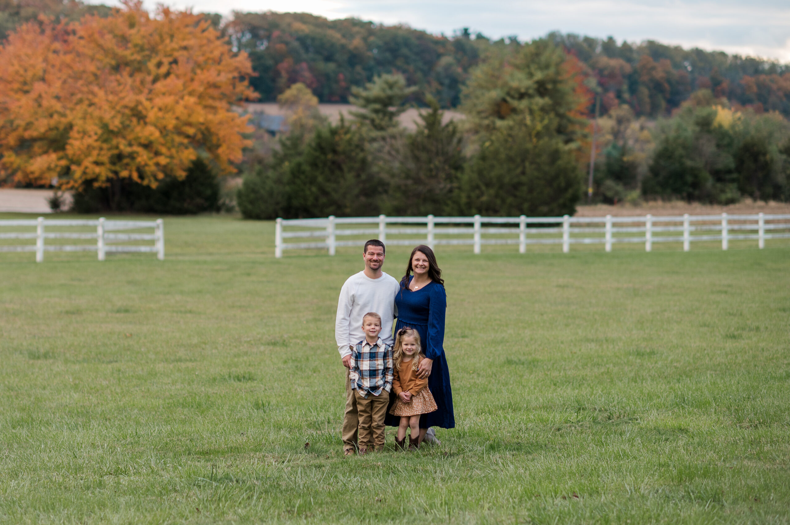Family of four standing together in a field surrounded by fall foliage during a fall mini session in York, PA.