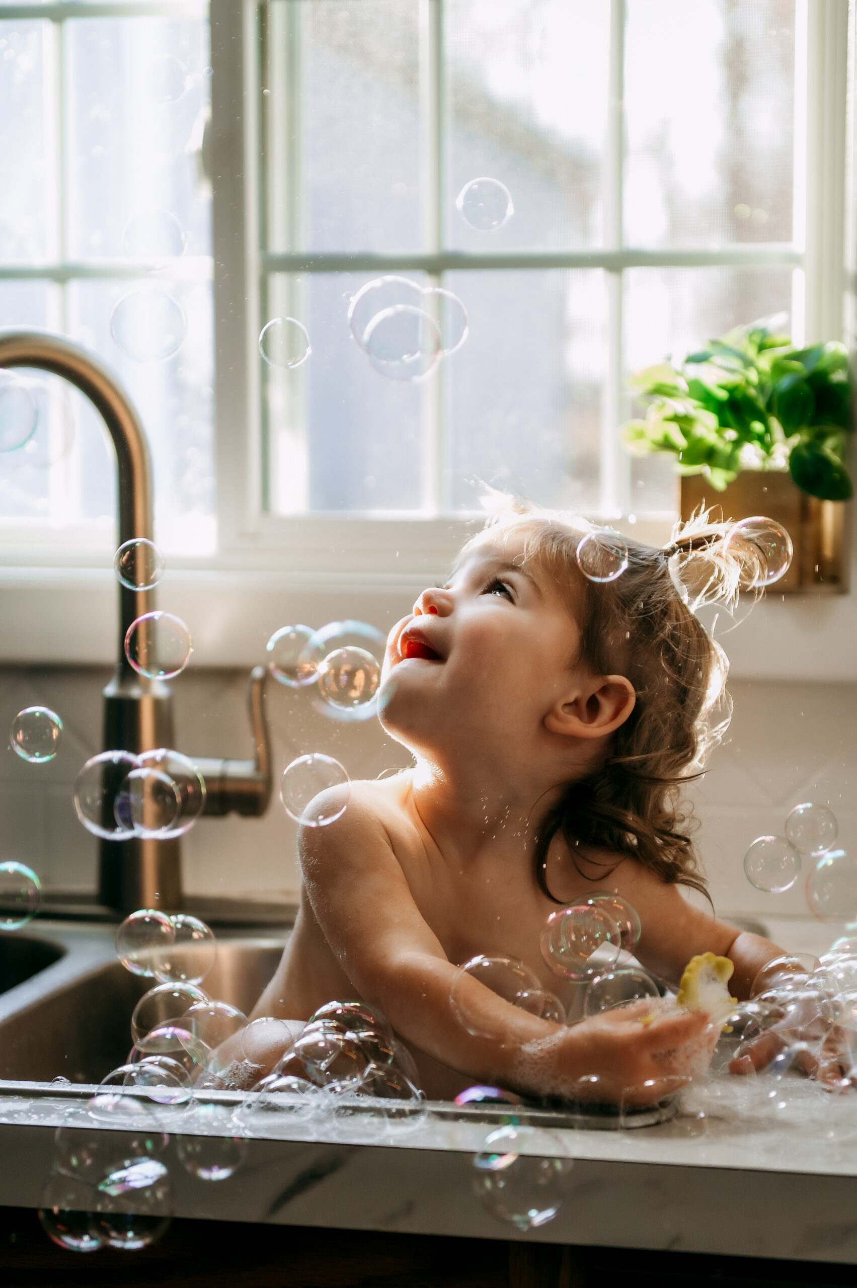 Young girl looking up at bubbles while playing in the kitchen sink during a documentary family session
